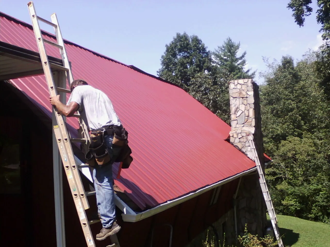 Roofer on a ladder beside a red metal roof and stone chimney — Soffit & Fascia Repair by Roofing Brothers in Marine On Saint Croix